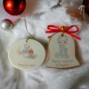 May include: Two porcelain Christmas ornaments featuring Precious Moments characters. One ornament is a round disc with a picture of two children, one in a blue hat and the other in a red hat, with the text "Pieces On Earth". The other ornament is a bell shape with a picture of a child in a blue outfit and the text "May Your Christmas Be Delightful".