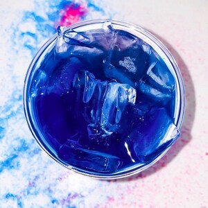 Può includere: Un bicchiere pieno di liquido blu e cubetti di ghiaccio. Il liquido è di un colore blu vibrante e i cubetti di ghiaccio sono parzialmente sciolti.