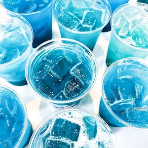 May include: An overhead shot of several clear plastic cups filled with a vibrant blue beverage and ice cubes. The drinks vary in shade from light turquoise to deep azure, creating a refreshing and cool visual.