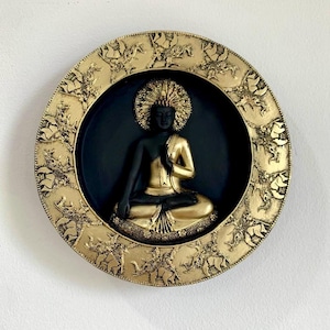 May include: A gold and black Buddha statue in a circular frame. The Buddha is seated in a meditative pose, with a gold robe and halo. The frame is gold with detailed relief carvings of figures and animals, creating a decorative wall hanging.