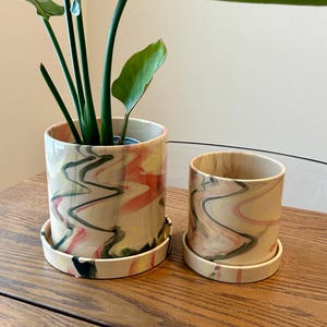 May include: Two ceramic plant pots with matching saucers. The pots have a beige base with a colorful, swirled design in shades of pink, yellow, and green. One pot is taller than the other. A green plant with large leaves is in the taller pot.