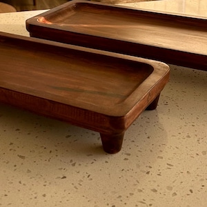 May include: Two rectangular wooden trays with rounded corners and short, rounded legs. The trays are a rich, dark brown color, and the wood grain is visible. The trays are empty and sit on a speckled, light-colored surface. The trays are likely for serving or display.