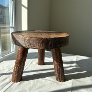 May include: A small, round wooden stool with three sturdy legs. The stool's top is a dark brown, circular piece of wood, and the legs are a similar, darker shade. The wood grain is visible, adding to the rustic aesthetic. This piece is suitable for home decor.