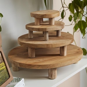 May include: A set of four round, light brown wooden risers of varying sizes stacked in a tiered formation. Each circular wooden platform features a smooth, natural grain finish and is supported by three short, sturdy cylindrical wooden legs.