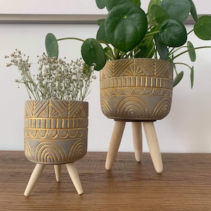 May include: Two gray and gold decorative planters with geometric patterns and wooden legs. One planter holds a green plant with round leaves, and the other holds dried white flowers. The planters are on a wooden surface.