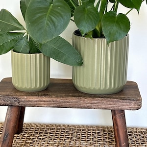 May include: Two green ribbed ceramic plant pots of different sizes, each holding a leafy green plant, sit on a small, dark brown wooden bench. The bench has four dark brown legs and is placed on a woven wicker basket.