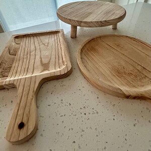 May include: A collection of wooden serving pieces, including a round cake stand with three legs, a rectangular tray with a handle, and an oval serving platter. The items are made of light-colored wood with visible grain.