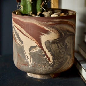 May include: A cylindrical planter featuring a marbled pattern of earthy brown, cream, and dark gray tones. The pot contains a green cactus with sharp spines and a layer of smooth, multi-colored river stones covering the soil surface.