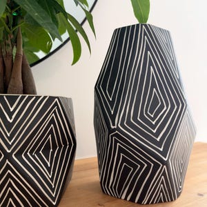 May include: Two black ceramic vessels with a faceted, geometric shape and white hand-painted linear patterns. The taller vessel stands upright, while the shorter vessel contains a green plant with multiple stems and leaves.