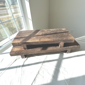 May include: A two-tiered wooden display stand featuring a dark brown stained finish with visible wood grain. The structure consists of a rectangular upper platform supported by short legs, resting on a wider, longer rectangular base platform.