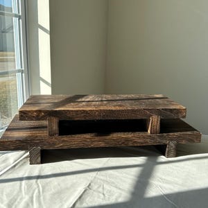 Könnte beinhalten: Ein zweistufiger Präsentationsständer aus Holz mit dunkelbraun gebeizter Oberfläche und sichtbarer Holzmaserung. Die Struktur besteht aus zwei rechteckigen Plattformen, die von kurzen, stabilen Holzbeinen getragen werden.