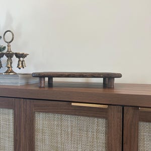 May include: A rectangular dark brown wooden riser with a smooth, flat top surface and four short, sturdy cylindrical legs. The wood grain is visible across the surface, and the edges of the riser are rounded, providing a stable base for display.