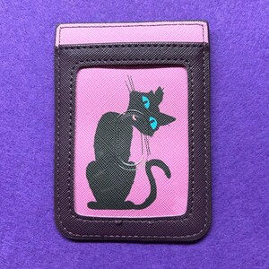 May include: A rectangular cardholder with a black cat illustration on a pink background. The cat has blue eyes and is set against a pink backdrop, framed by a dark brown border and a pink top.