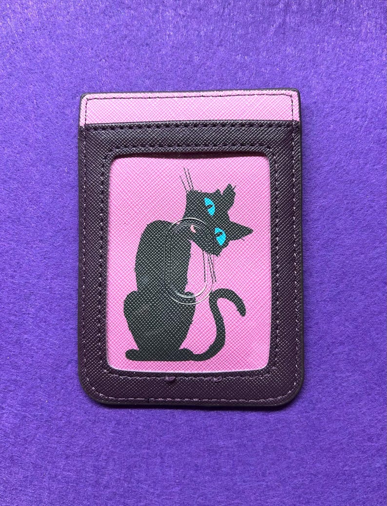 May include: A rectangular cardholder with a black cat illustration on a pink background. The cat has blue eyes and is set against a pink backdrop, framed by a dark brown border and a pink top.