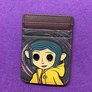 May include: A rectangular card holder featuring a cartoon character with blue hair and button eyes, wearing a yellow jacket. The card holder has a dark brown top section with card slots and a colorful swirling background.