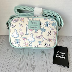 Puede incluir: Un bolso bandolera rectangular con una correa y ribetes azul claro. El bolso presenta un fondo crema con un patrón repetido de ilustraciones temáticas de Alicia en el país de las maravillas en azul, rosa y blanco. Una etiqueta negra con el logotipo de Disney Loungefly está adjunta.