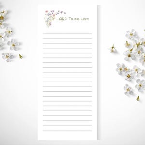 May include: A white rectangular notepad with horizontal black lines for writing. The top left corner features a delicate watercolor illustration of small purple and pink wildflowers. The text Ally's To Do List: is printed in a dark script font.
