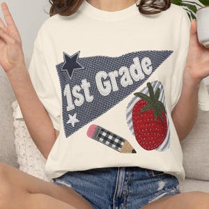 Peut inclure: T-shirt crème avec un motif graphique coloré pour la première année. Le motif comprend un fanion bleu avec le texte "1st Grade", une fraise rouge et un crayon. Le t-shirt est associé à un short en jean.