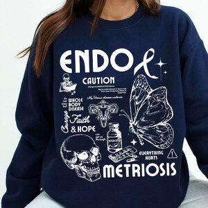 May include: Navy blue sweatshirt with a white graphic design that says "ENDO & METRIOSIS" with a skull, butterfly, and other medical illustrations. The text "CAUTION" is also included in the design.