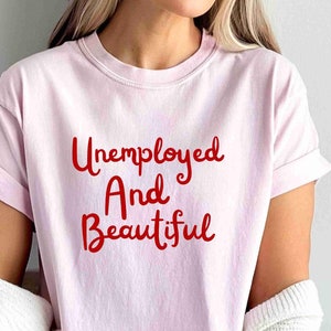 May include: A light pink t-shirt with the words "Unemployed And Beautiful" in red script. The shirt has short sleeves and a crew neck. The shirt is made of a soft material.