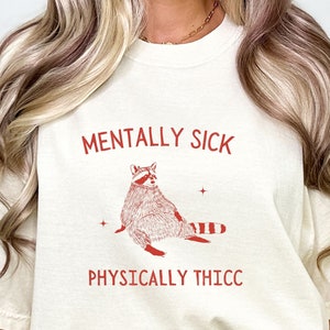 May include: A white t-shirt with red text that reads "Mentally Sick" and "Physically Thicc". A cartoon raccoon is printed in red on the shirt.