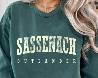 May include: A dark green sweatshirt with the text "Sassenach Outlander" printed in a vintage-style font in cream.