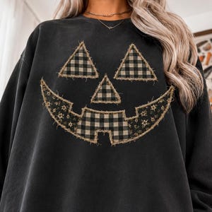 May include: A charcoal gray long-sleeve sweatshirt featuring a large jack-o'-lantern face graphic. The eyes, nose, and mouth are constructed from frayed-edge fabric patches in black and white buffalo plaid and a dark floral pattern.