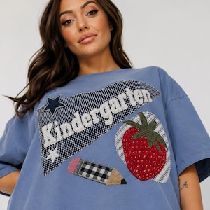 May include: A blue short-sleeve t-shirt featuring a large applique patch with the word Kindergarten in white text. The patch includes a black and white gingham pennant, a red strawberry with a green stem, and a pencil with a gingham pattern.