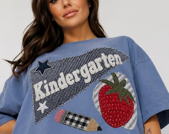 May include: A blue short-sleeve t-shirt featuring a large applique patch with the word Kindergarten in white text. The patch includes a black and white gingham pennant, a red strawberry with a green stem, and a pencil with a gingham pattern.