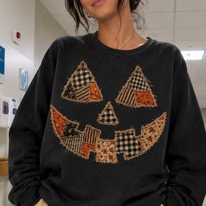 May include: A black crewneck sweatshirt featuring a large jack-o'-lantern face graphic on the front. The pumpkin face is constructed from various fabric patches in orange, black, and white with plaid, floral, and striped patterns and frayed edges.