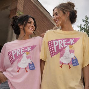 May include: A light pink t-shirt and a pale yellow t-shirt, each featuring a graphic of a white goose carrying a blue bag labeled "We Rock" and a pink gingham pennant with the text "PRE-K" in white letters, paired with gray denim jeans.