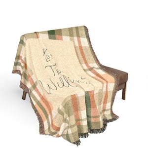 May include: A woven throw blanket featuring a plaid pattern in shades of green, orange, and cream. The center displays a cream-colored panel with a line-art drawing of a house and the text "The Willins" written in a cursive black script font.