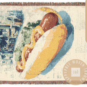 May include: Woven throw blanket featuring a colorful illustration of a hot dog with mustard. The blanket has a cream-colored background with a fringe border. The text "HAPPYPRET WOVEN THROWS" is in a circle on the bottom right.