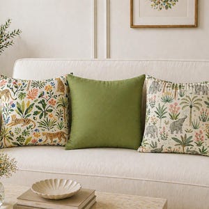 May include: Three decorative cushions on a white sofa. Two cushions feature colourful botanical and animal prints, including leopards and elephants. The centre cushion is a solid olive green. The cushions add a touch of nature-inspired decor.