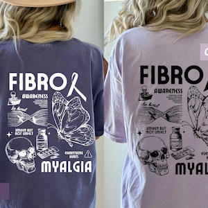 May include: Two t-shirts with a graphic design featuring a butterfly, a skull, pills, and the text "Fibro Awareness" and "Everything Hurts Myalgia". The t-shirt on the left is a dark purple color and the t-shirt on the right is a light purple color.