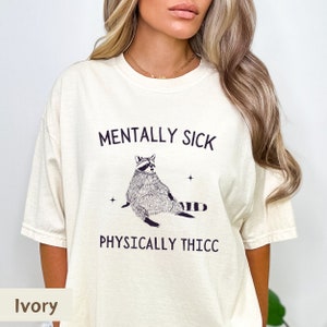 May include: A white t-shirt with the text "Mentally Sick" and "Physically Thicc" printed on it. A cartoon raccoon is sitting in between the text. The t-shirt is available in ivory.