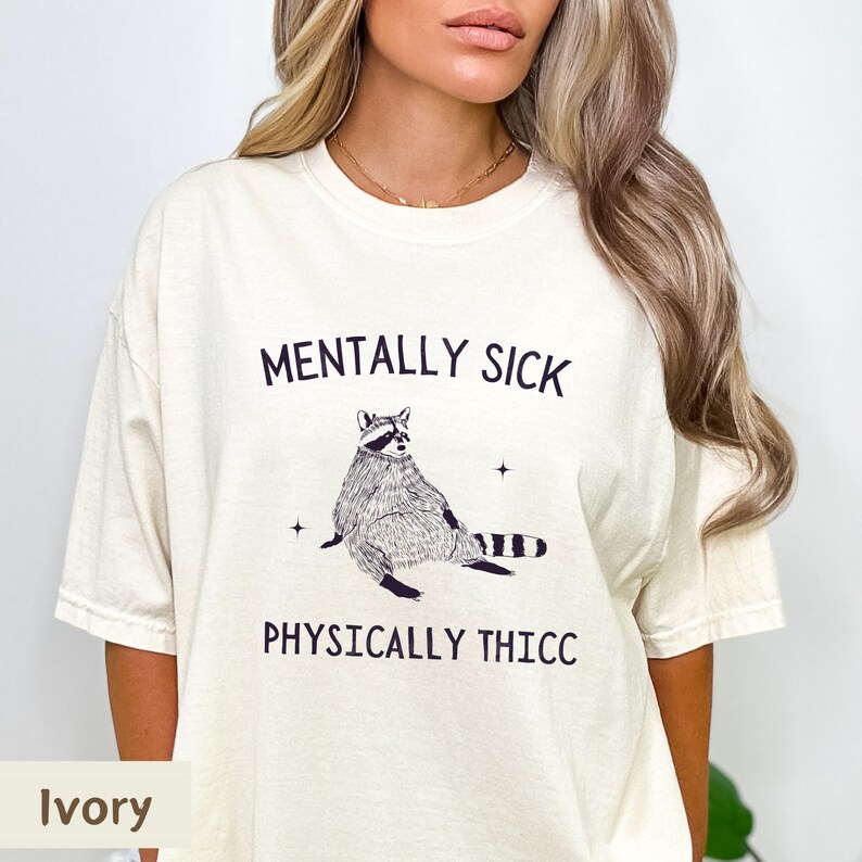 May include: A white t-shirt with the text "Mentally Sick" and "Physically Thicc" printed on it. A cartoon raccoon is sitting in between the text. The t-shirt is available in ivory.