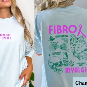 May include: Light blue t-shirt with a pink ribbon and green graphic design. The graphic includes a skull, pills, a butterfly, and the text "Fibro Awareness" and "Myalgia".