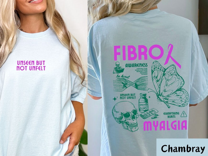 May include: Light blue t-shirt with a pink ribbon and green graphic design. The graphic includes a skull, pills, a butterfly, and the text "Fibro Awareness" and "Myalgia".