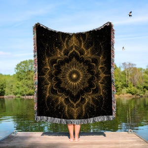 May include: Black throw blanket with a gold mandala design. The blanket has a fringed edge with multiple colors. The mandala design is a detailed, symmetrical pattern with a central point and radiating elements.