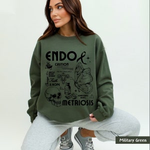 May include: A green sweatshirt with a black graphic design that says "ENDO CAUTION" and includes a butterfly, skull, and other medical symbols. The text "EVERYTHING HURTS" and "METRIOSIS" are also included in the design.