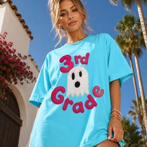 Op de afbeelding: Een fel turquoise T-shirt met korte mouwen en een centrale afbeelding met de tekst 3rd Grade in dikke roze letters. Tussen de woorden staat een wit spookfiguur met zwarte ogen en een zwarte mond, uitgevoerd in een textuurrijke applicatiestijl.