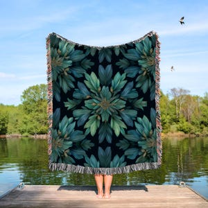 May include: A dark blanket with a symmetrical floral design in shades of teal and green. The blanket has a black background and fringed edges with tan, orange, and white threads. The blanket is held up by a person standing on a wooden dock, with a lake and trees in the background.