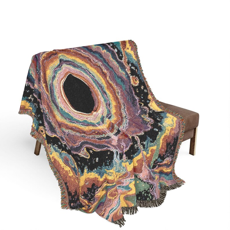 Puede incluir: Una manta tejida que presenta un gran patr&oacute;n central inspirado en una geoda con anillos conc&eacute;ntricos de color negro, morado oscuro, amarillo dorado y azul verdoso. El dise&ntilde;o imita formaciones minerales naturales con un borde con flecos.