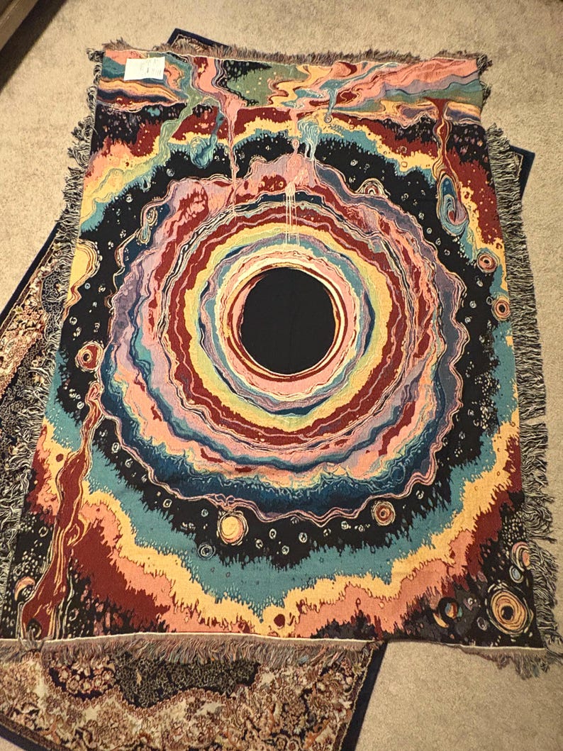 Puede incluir: Una manta tejida rectangular que presenta un gran c&iacute;rculo negro central rodeado por anillos conc&eacute;ntricos de colores arremolinados que incluyen rosa, amarillo, azul verdoso y rojo oscuro. El dise&ntilde;o incluye peque&ntilde;os motivos circulares y flecos.