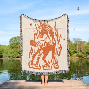 May include: A beige throw blanket with a burnt orange horse and flame design. The blanket has a fringed edge with multiple colors. The Chinese character for "blessing" is in the upper left corner. The blanket is held up by a person on a wooden dock.