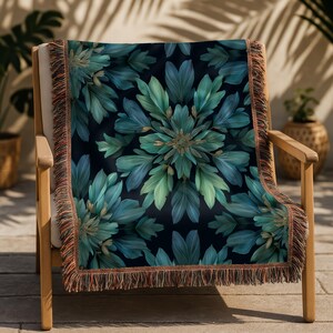 May include: A decorative throw blanket with a dark background and a symmetrical floral pattern in shades of teal and green. The blanket has a fringed edge in shades of brown and orange. The blanket is draped over a wooden chair.