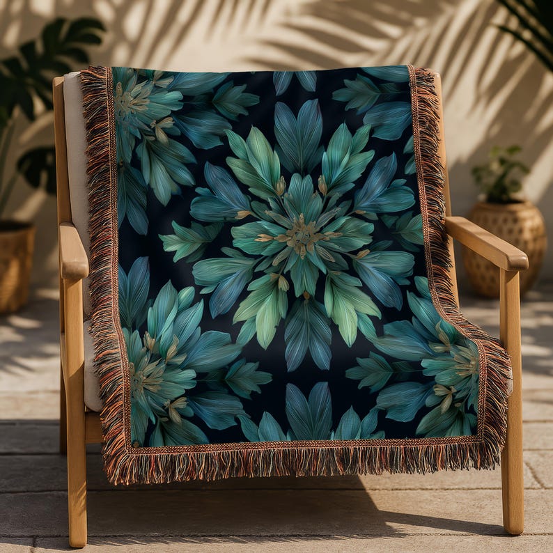 May include: A decorative throw blanket with a dark background and a symmetrical floral pattern in shades of teal and green. The blanket has a fringed edge in shades of brown and orange. The blanket is draped over a wooden chair.