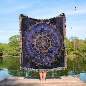 May include: A large, rectangular throw blanket with a mandala design. The blanket features a dark blue background with a gold mandala pattern in the center. The edges have a fringe in shades of brown, tan, and white. The blanket is held up against a blue sky.