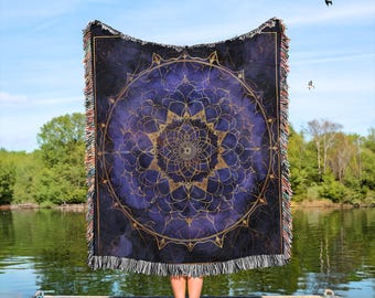 May include: A large, rectangular throw blanket with a mandala design. The blanket features a dark blue background with a gold mandala pattern in the center. The edges have a fringe in shades of brown, tan, and white. The blanket is held up against a blue sky.